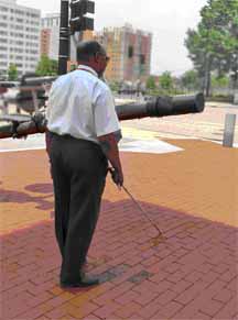 photo shows a man using a white cane and walking in a plaza toward a large rod that is extended diagonally across his path; if he continues walking, his cane will go under the rod and he will bump into it with his chest.