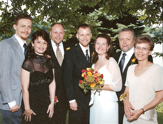 Standing in front of trees and smiling are Stephan with a mustache, goatee and short curly brown hair, Jomania with a black dress with black lace around the neck and shoulders, Paul with a goatee, Mark the groom wearing a dark suit and tie with Jill holding flowers and wearing a white wedding dress with straps and an opaque white shawl or jacket, Fred and Dona.  All men have suits and ties, and Dona is wearing a cream-colored scoop-necked short-sleeved dress.