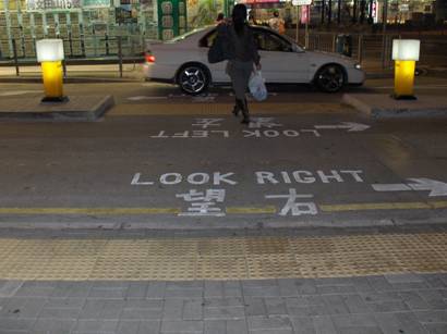 photo shows a mid-block crosswalk where there is no curb anywhere along the entire width of the crosswalk (about 15 feet wide). Along the edge of the street are rows of yellow standard detectable warning tiles (tiles with rows of flattened domes).  Written in the street in English and Chinese is 'look right' with an arrow pointing to the right.