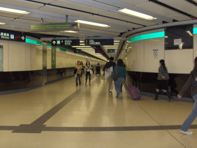 Photo shows a busy hallway with a person in a wheelchair traveling away from us, far away in the hall.  On the floor along the middle of the hall is a tactile strip about a foot wide, intersecting with the strip of another hall.