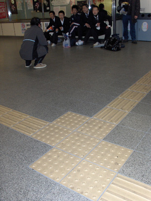 Photo shows two strips on the floor, each about a foot wide and intersecting to form overlapping squares each about 2 feet wide.  There are 4 raised bars extending along the strips, and in the square where they intersect are flattened domes in rows about an inch apart (standard detectable warning surface).