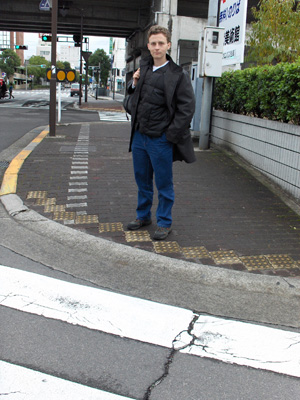 photo shows Stephan standing at a rounded corner (with a wide radius) which blends into the street.  Running along the sidewalk from the previous corner is a row of tactile tiles, about a foot wide and placed about foot apart.  Running along the edge of the street are more one-foot tiles that are aligned with the crosswalk, most of them have one corner touching the corner of the next tile, looking like a stairway.