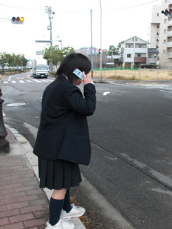 a schoolgirl in uniform stands at the edge of a street using her cell phone.  The brick sidewalk is lowered at the crosswalk to be flush with the street, with a concrete border at the edge.