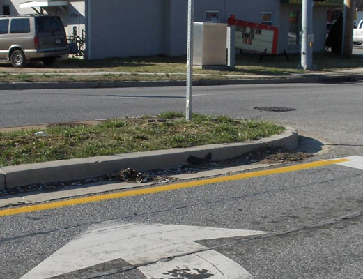 Photo shows a street with two grey parallel lines in the middle of the left-turning lane -- the lines are parallel and about 3 feet apart; a pedestrian button on a pole near the sidewalk and a large grey metal box sits in the grass on the other side of the sidewalk -- it is about 4 feet high, 2 feet deep, and 3 feet wide.