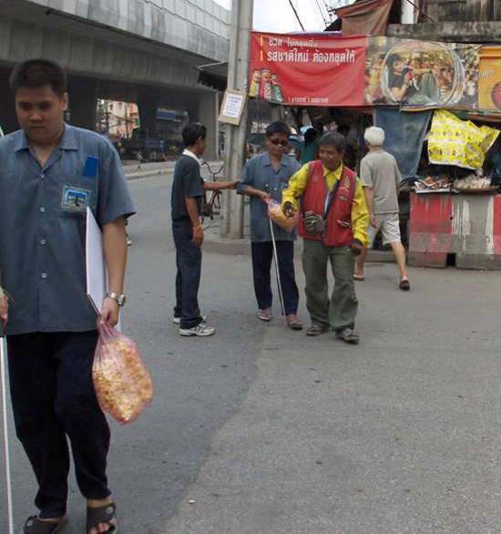 Photo shows both students crossing a quiet two-lane street with no traffic, coming from a sidewalk cluttered with vending stands and banners.  Both students are using canes and carrying a large bag of popcorn, one has a mailing envelope tucked under his arm.  One student is walking by himself and approaching close to the camera, the other is about 10 or 15 feet behind and is approached by two people, one of whom is wearing a yellow jacket and offering his arm on the student's left side, the other is on the student's right and reaching out to hold his right arm.