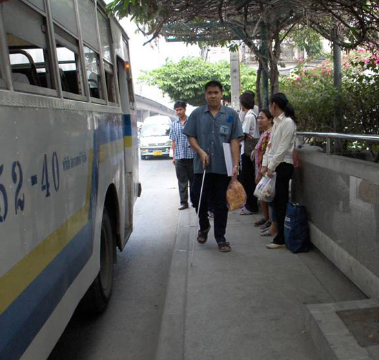 Photo shows the first student walking along the sidewalk with a bus on his right and people waiting along a railing on his left.  The other student is behind him but is not visible in the photo.