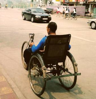 photo shows the back of a man in a chair at the left side of the street.  The chair has three wheels that look like bicyle wheels about 2.5 feet in diameter - one wheel on each side of the chair and a third in the front.  His left hand is resting on the chair and we can't see his right hand.