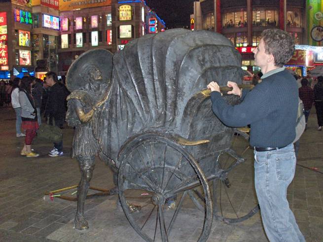 Stephan pushes the back handle of a bronze statue of a rickshaw with a bronze man on the other side.