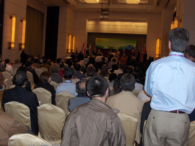 two photos show a large room with standing room only, and an empty stage flanked with flags.