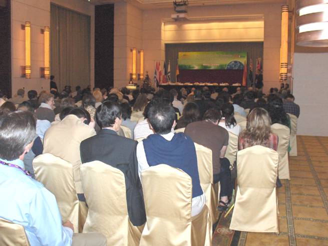 two photos show a large room with standing room only, and an empty stage flanked with flags.