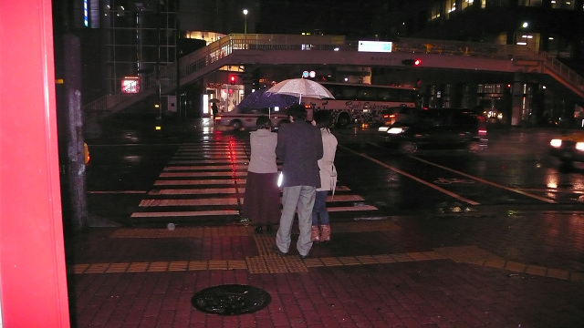Photos show Dona holding an umbrella and standing at a crosswalk at night.  The crosswalk has white lines running left-to-right, and along the middle of the lines going across the street is a dark line which is the guiding strip.