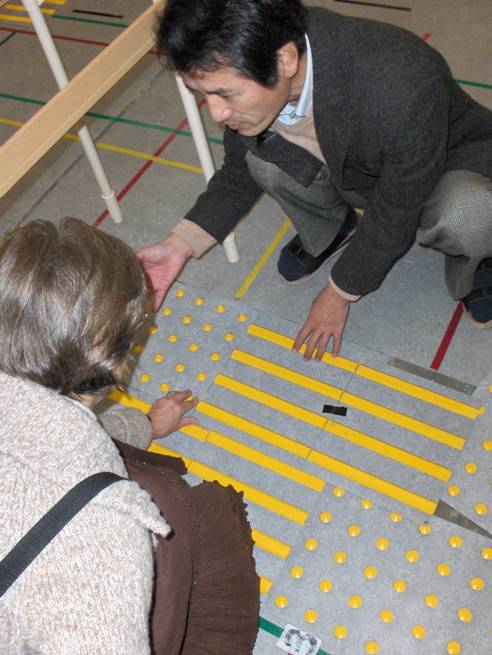 These 3 photos show Dr. Nakamura and Dona kneeling on the ground looking at about a half dozen black mats, each about 1'x2' with raised yellow lines or circles.  They are next to two parallel bars supported about waist-high, where subjects can walk between the bars.