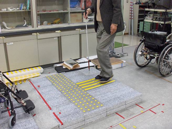 Photo shows Professor Nakamura standing on the raised yellow lines.  In front of the lines are mats with raised circles.