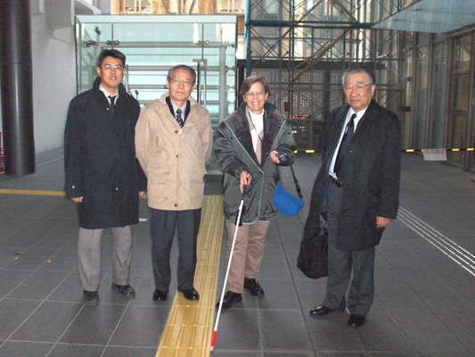 photo shows the three researchers and Dona, standing and smiling in a large gymnasium-sized room with a row of tactile bars on the floor.  Dona is holding a white cane in one hand and in the other is a device about 6 inches long.