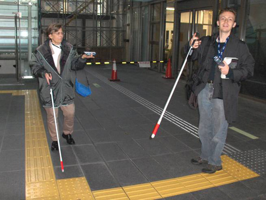 Dona and Stephan stand along the tactile rows in the large room, each holding a white cane in one hand and a device about 6 inches long in the other.