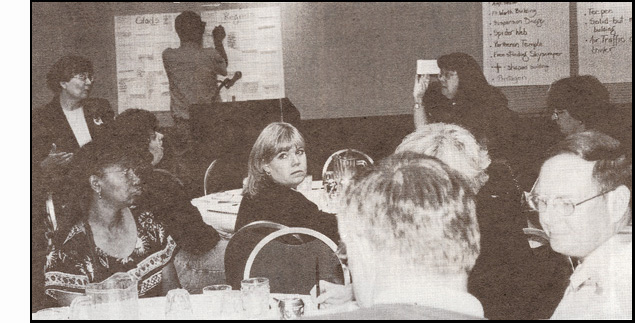 Photo shows people seated at several tables, some are writing, others looking at other people and one is looking back toward the camera.  Mary Nelle is standing in front and looking toward Annette Skellenger who is walking toward Mary Nelle while talking and holding up a card.  On the front wall are large sheets of paper and Lukas Franck is writing on one of them.