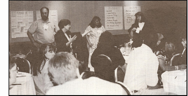 Photo shows the same people seated at the tables, most are leaning forward toward the front of the room.  Four people are standing in front of the room:  Mary Nelle is looking toward Annette Skellenger and speaking, Annette is standing with a card and watching Mary Nelle, Lukas Franck is standing in front of the sheets of paper posted on the wall, with hands on hips.