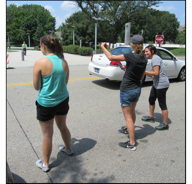 Photo shows 3 women standing at the edge of a two-lane street as a car passes them.