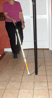 Series of photos shows a woman approaching a pole, holding a cane in her right hand which is centered in front of her.  The cane tip starts out on the woman's left and as she moves forward, the tip moves to the woman's right, barely missing the pole and reaches to the right of the pole.  When the tip is moved back to the left, the shaft of the cane contacts the pole.