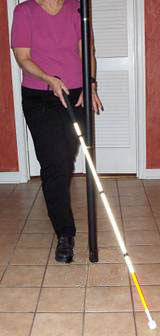 Series of photos shows a woman approaching a pole, holding a cane in her right hand which is centered in front of her.  The cane tip starts out on the woman's left and as she moves forward, the tip moves to the woman's right, barely missing the pole and reaches to the right of the pole.  When the tip is moved back to the left, the shaft of the cane contacts the pole.
