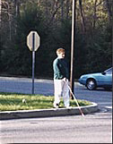 3 photos show a boy with a white cane stepping off a curb, walking across the street and reaching the other side of the street -- his cane has crossed the white line at the edge of the street's shoulder.