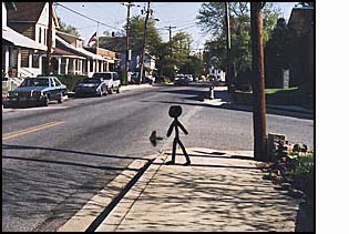 A stick-figure/person stands on a corner facing a two-lane street with cars parked on the other side and small houses close together.  The street goes slightly downhill to the right.