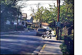 The figure/person on the corner faces a street with two moving lanes plus enough room for cars to park on both sides.  To the right of the figure (north) the street is straight for about a block, and has a minivan and other vehicles parked along the street, close to the corner.
