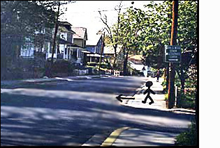 The figure/person on the corner faces a street with two moving lanes plus enough room for cars to park on both sides.  To the right of the figure (north) the street is straight for about a block. There are no cars parked in the street.