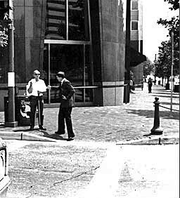 Picture shows a deaf-blind man wearing a white short-sleeved shirt and dark pants and sunglasses standing at a crosswalk facing the street, holding up a card with his right hand and holding a white cane in front of him with his left hand.  He is standing in front of some newspaper boxes and beside a telephone pole.  Another man wearing a suit and tie is approaching him from the street and reaching out to touch his left elbow.