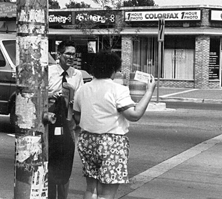 This series of 3 photos shows a deaf-blind woman wearing short-sleeved white shirt and flowered shorts.  We see her from the back as she stands at the crosswalk holding up a card in her right hand.  A man with white shirt and tie, holding his business coat over his shoulder, crosses the street toward her and then stands in the street to her left, looking at her card.  Last photo shows them standing at the curb waiting to cross, she is holding his left arm and he looks toward her.