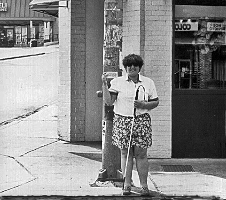 Photo shows deaf-blind woman wearing short-sleeved white shirt and flowered shorts standing at the curb and smiling broadly, holding up a card in front of her right shoulder and holding her white cane with the left hand.