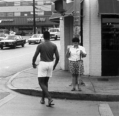 This series of 4 photos were taken from the middle of the street.  The deaf-blind woman faces the street and holds up the card with her right hand, while a man in the crosswalk, wearing a dark tank-top, white shorts and flip-flops, walks toward the curb at her right, turning his head to look at the card.  Then he stands in the street next to the curb, leaning forward to look at the card while she stands and smiles.  Last photo they walk into the street while she holds his arm, both smiling and looking forward.