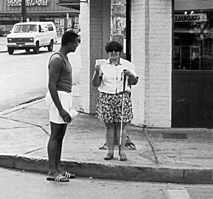 This series of 3 photos were taken from the middle of the street.  The deaf-blind woman faces the street and holds up the card with her right hand, while a man in the crosswalk, wearing a dark tank-top, white shorts and flip-flops, walks toward the curb at her right, turning his head to look at the card.  Then he stands in the street next to the curb, leaning forward to look at the card while she stands and smiles.  Last photo they walk into the street while she holds his arm, both smiling and looking forward.