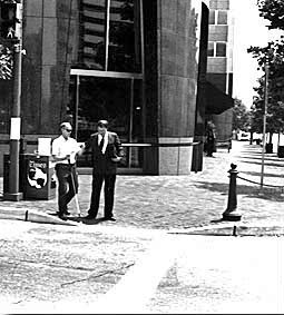 Picture shows the same two men as the last photo.  They are both facing the street and the deaf-blind man with the white shirt is starting to walk, holding his white cane in his right hand and reaching for the other man with his left hand.  The man in the suit is beside him and holding his left elbow, looking down. 