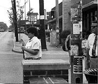 Photo shows a woman wearing sunglasses and a white short-sleeved blouse, standing at the curb facing the street and smiling, holding a white cane in her right hand and her left hand is holding up a card in front of her shoulder.  To  her left is a wall that obscures her from our view from the waist down, and behind that is a telephone pole.  On  the other side of the pole is a man wearing a shirt and tie and sunglasses, stopping to look at her.