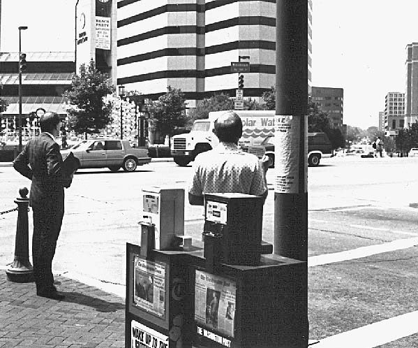 photo shows two men standing at a crosswalk facing the street -- we are standing behind them. The man on the left is wearing a suit and holding a newspaper.  He  is looking to his left into the street.  The  other man is about 10 feet to his right, wearing a white short-sleeved shirt and holding up a card in his right hand.  Behind  and to his right are magazine boxes and a telephone pole, which makes us unable to see anything except his back and head.