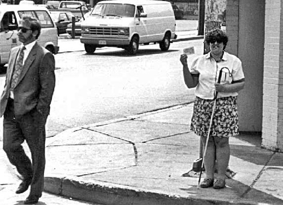 We are standing in the street looking at the deaf-blind woman who is smiling and holding up the card with her right hand and her cane is in her left hand.  About 5 feet to her right is a bearded man wearing a business suit and sunglasses, hands in his pockets, looking forward and stepping out into the street.