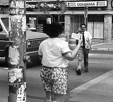 This series of 4 photos shows a deaf-blind woman wearing short-sleeved white shirt and flowered shorts.  We see her from the back as she stands at the crosswalk holding up a card in her right hand.  A man with white shirt and tie, holding his business coat over his shoulder, crosses the street toward her and then stands in the street to her left, looking at her card.  Last photo shows them standing at the curb waiting to cross, she is holding his left arm and he looks toward her.