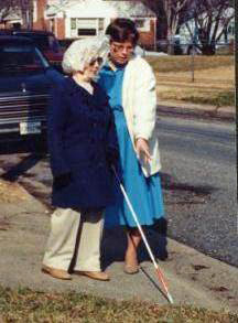 An elderly woman with a white cane has veered toward the street in a driveway; Dona stands next to her and points out the slope and grass.