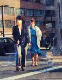 A young woman with a cane walks across a street, while Dona walks about 10 feet behind.