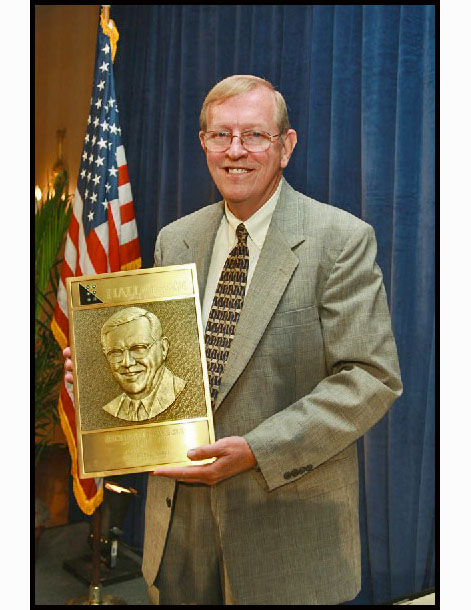 Photo shows Rick in front of a blue curtain and an American flag, holding his APH Hall of Fame plaque.