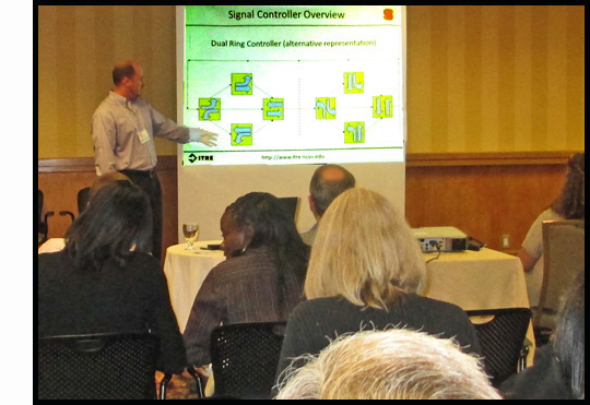 Chris is standing on front of a room full of participants and points to diagrams on a powerpoint titled 'Signal Controler Overview.'