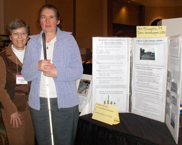 Photo shows Ann holding her cane with Dona standing next to a table with a folded display about 3 feet high and 4 feet wide.  The display has the text and photographs from this page.