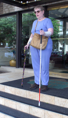 The woman is standing on the top of the stairs, with her support cane beside her.  The long white cane is touching the top of the next step.