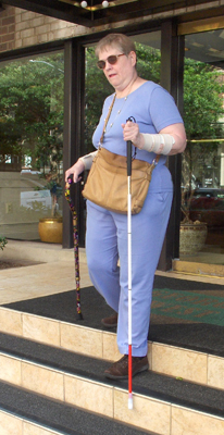 The woman's left foot is now on the next step, her right foot and her support cane are still on the top step.  The long white cane is reaching over the edge of the step that her left foot is standing on.