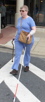 Photo shows a woman entering a crosswalk.  She has ace bandages around both of her wrists, and is supporting herself with a support cane in her right hand close to her body, and reaching a long white cane ahead of her with her left hand (her right foot is forward, and the tip of the long cane is far in front of her left foot).