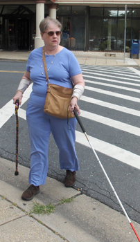 The woman has completed the crossing and is stepping out of the crosswalk.  She is still supporting herself with a support cane with a curved handle in her right hand, and reaching the long white cane ahead of her with her left hand (again her right foot is forward, and the tip of the long cane is reaching far ahead to her left).