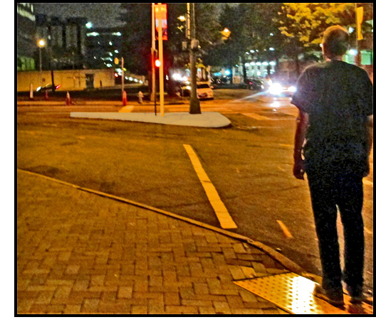 photo shows a pedestrian standing at the end of a channelized right-turning lane, facing the island and the crosswalk that leads to it.  The curbramp with detectable warnings are parallel to the curb, which has turned so much that it is almost parallel to the crosswalk and faces out into the intersection.