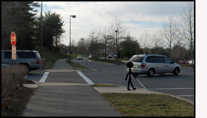 Photo shows a stick figure on a corner, facing to cross a 2-lane street that has no stop sign or traffic signal.  In the street beside the figure is a car at a stop sign, waiting to cross the same street.