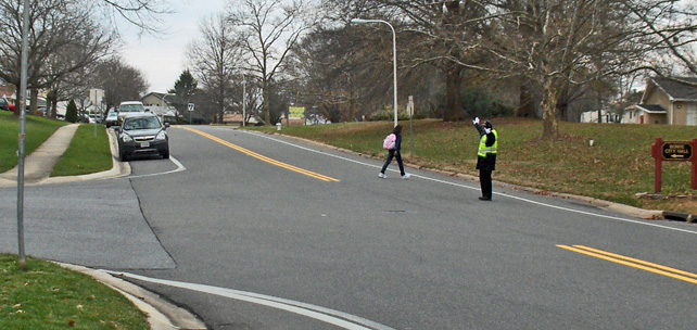 photo shows a T-intersection of two 2-lane streets.  A child is crossing the main street, with a crossing guard nearby.  The only cars visible on either street are parked.
